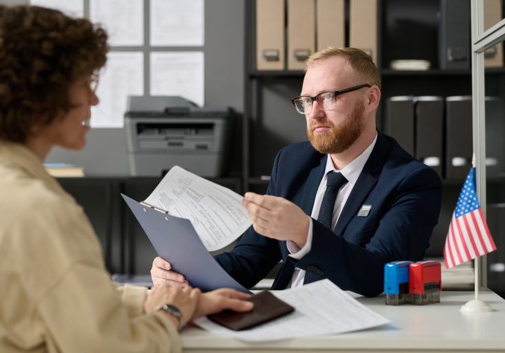 Portrait of bearded male consultant working in US embassy office and holding application form while talking to woman